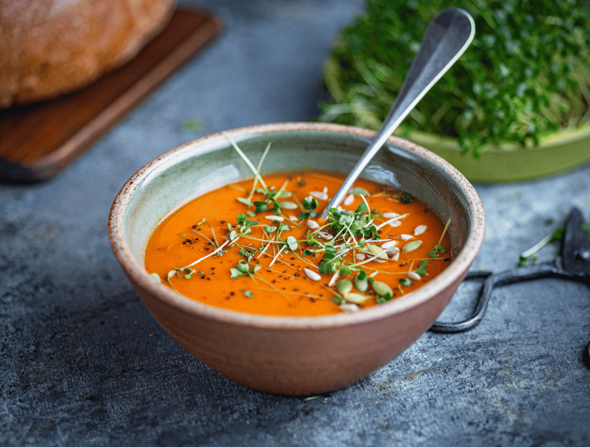 Bowl of soup with a spoon on a gray surface, surrounded by bread and greens.
