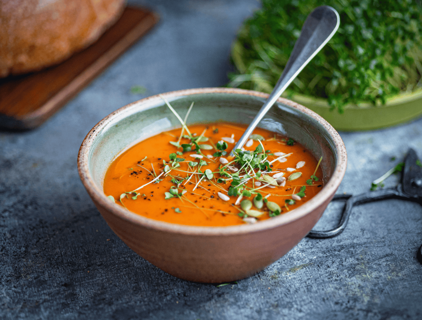 Bowl of soup with a spoon on a gray surface, surrounded by bread and greens.